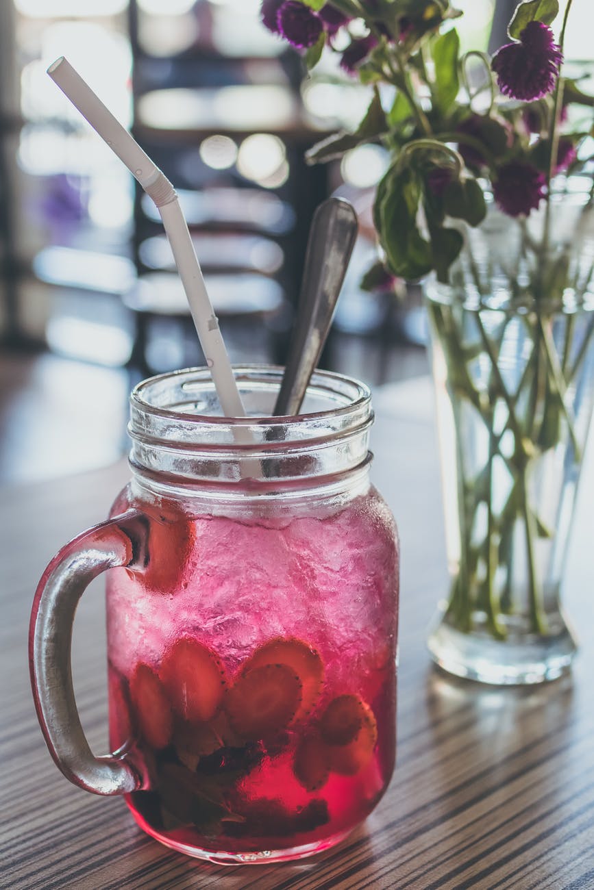 red beverage filled clear glass mason mug with straw beside purple flowers in vase